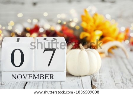 White wood calendar blocks with the date November 7th and autumn decorations over a wooden table. Selective focus with blurred background. 