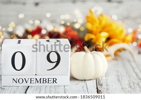 White wood calendar blocks with the date November 9th and autumn decorations over a wooden table. Selective focus with blurred background. 
