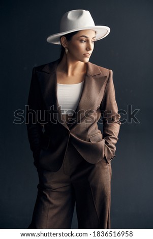 portrait photo on a dark background of a beautiful stylish girl in a brown suit, white top and white hat, she stands and looks away 