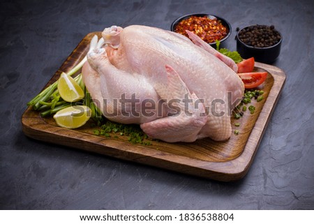 Raw whole chicken with skin arranged on wooden board and garnished with parsely,small tomato,spring onion,chilli flakes and lemon slices with stone textured colour as background ,isolated Royalty-Free Stock Photo #1836538804