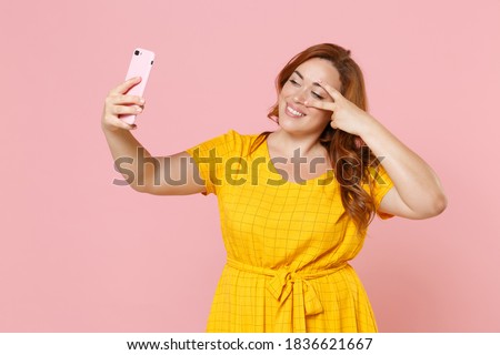 Fun young redhead plus size body positive chubby overweight woman in yellow dress posing doing selfie shot on mobile phone showing victory sign isolated on pastel pink color background studio portrait