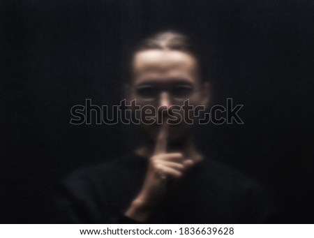 Blurred scary man silhouette behind a transparent plastic. Man in a black clothes and glasses gesturing silence.