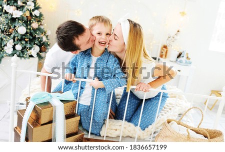 young family - mom, dad and little son in pajamas sitting on the bed against the background of a Christmas tree with gifts hugging and smiling.