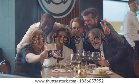 Happy diverse friends having dinner and taking selfie on smartphone at restaurant. Cheerful multiethnic group of people dining together and taking photo on cellphone in modern cafe