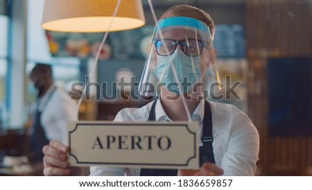 Waiter in safety mask and shield checking aperto hanging sign in small italian cafe. Small business owner wearing apron and protective mask turning open closed sign written in italian