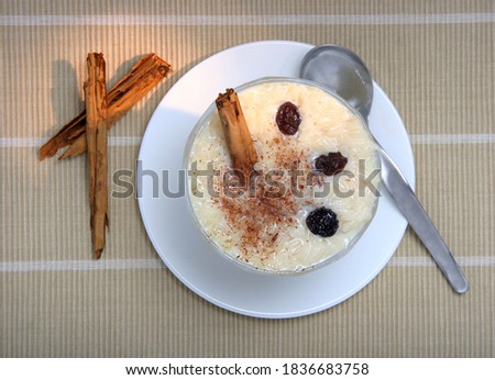 Sweet rice pudding in a bowl, with cinnamon powder and raisins on top, with some cinnamon sticks and a spoon on the side. All on a place mate on a table. Royalty-Free Stock Photo #1836683758