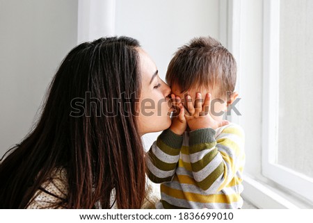 Portrait of young mother kissing her little son in forehead at home. Beautiful female with straight brunette hair playing with child at living room. Close up, copy space, background. Royalty-Free Stock Photo #1836699010