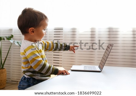 Two year old boy staring at the laptop screen with fascinated facial expression. Toddler boy sitting by the table at home with notebook computer. Close up, copy space, background.