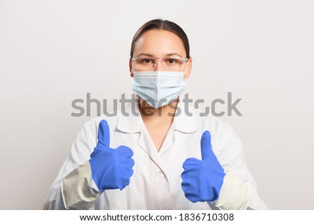 Young woman doctor in protective mask and gloves shows thumb up gesture.