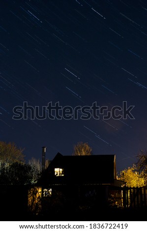 Beautiful star trail image during the night of the meteor shower in the summer. Night landscape with a country house with glowing windows. Magic nature. Astromomical phenomenon