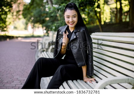 Portrat of young pretty laughing Asian woman in fashionable black pants and leather jacket, sitting on the bench green city park background. Lifestyle, leisure and people concept.