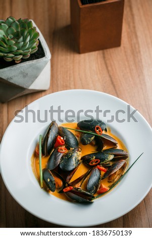 Gourmet serving of spicy tomato soup with fried mussels and chili peppers on a plate and wooden table isolated. Delicious seafood mussels . Lemon and baguette . Clams in the shells. Top view.