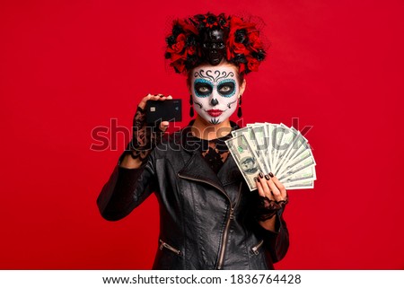 Scary young creepy lady calavera. wears artistic make-up for the feast of all the dead.Wears black leather jacket and lace gloves holds a pack of money and creadit card in hands, isolated in red