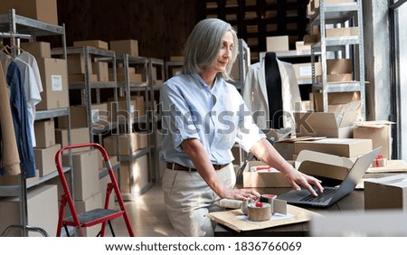 Older middle aged business woman entrepreneur, fashion clothing seller using laptop checking ecommerce dropshipping order packing online shop shipping delivery parcels boxes at workplace in warehouse. Royalty-Free Stock Photo #1836766069