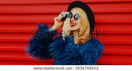 Portrait of young woman with retro camera taking picture wearing blue faux fur coat, round hat over red background
