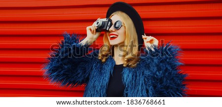 Stylish smiling young woman with retro camera taking picture wearing blue faux fur coat, round hat over red background