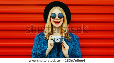 Portrait of happy smiling woman with retro camera wearing blue faux fur coat, round hat over red background