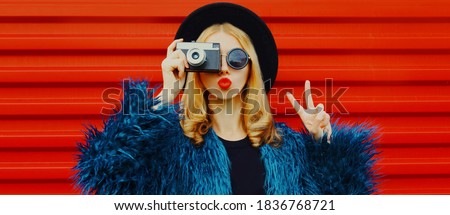 Fashion portrait of woman with retro camera taking picture blowing red lips sending sweet air kiss wearing blue faux fur coat over red background