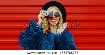 Portrait of surprised woman with retro camera taking picture wearing blue faux fur coat, round hat over red background