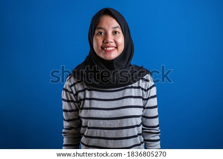 Muslim college student girl smiles to camera against blue background with confident gesture.