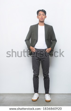 full length young business man in wearing black suit ,white t-shirt with black pants and sneakers shoes hands in pockets posing on gray background