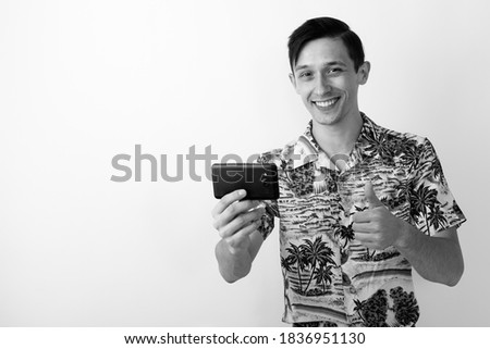 Studio shot of young happy tourist man giving thumb up and taking picture with phone
