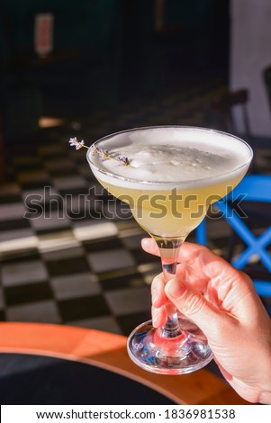 Summer cocktail in a Martini glass with fresh lavender flower. Lavander alcohol drink, refreshing beverage in restaurant. In womans hand over blurred restaurant background.
