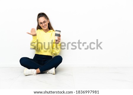Young woman sitting on the floor holding a take away coffee inviting to come with hand. Happy that you came