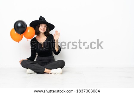 Young witch holding black and orange air balloons sitting on the floor saluting with hand with happy expression