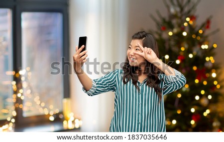 winter holidays and people concept - happy smiling young asian woman taking selfie by smartphone showing peace hand sign over christmas tree on background