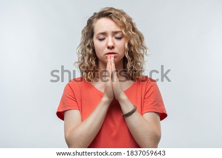 Portrait of joyous blonde girl with curly hair in casual t shirt keeping palms together and praying. She meditating, praying for peace and love, having calm and peaceful facial expression