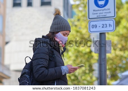 Woman in a mask on the background of a sign that warns people to cover mouth and nose on the Marienplatz (Mary's Square) in Munich, Germany. Warmly dressed woman in a mask is holding her phone Royalty-Free Stock Photo #1837081243