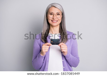 Photo portrait of old woman holding credit card with two hands wearing violet cardigan isolated on grey colored background