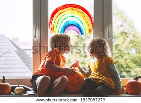 Preschool children on background of painting rainbow on window.  Family preparing for celebrating Halloween during quarantine Pandemic Coronavirus Covid-19 at home. Kids leisure activities indoors.