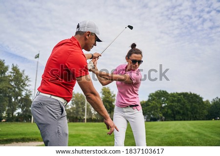 Skilled middle-aged male instructor in a cap explaining the golf fundamentals to a female beginner Royalty-Free Stock Photo #1837103617