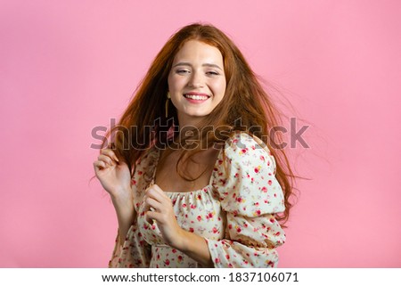 Young woman with long red wavy and shiny hairstyle looking to camera, flirting. Beautiful model girl demonstrate her elegant volume hair. Pink studio background.