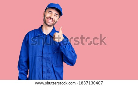 Young hispanic man wearing painter uniform smiling happy and positive, thumb up doing excellent and approval sign 