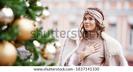 Outdoor portrait of young beautiful dreaming girl posing on street against background of decorated fir  tree.  Model wearing stylish warm clothes. Christmas, new year, winter holidays concept.  Banner