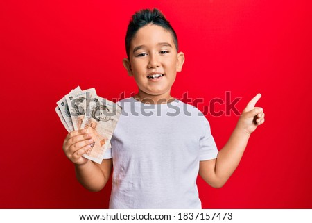 Little boy hispanic kid holding 10 united kingdom pounds banknotes smiling happy pointing with hand and finger to the side 
