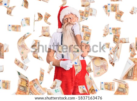Old senior man with grey hair and long beard wearing santa claus costume with suspenders serious face thinking about question with hand on chin, thoughtful about confusing idea