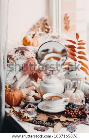 A cup of hot tea with lemon in an early autumn morning by the window. A stack of sweaters, yellow leaves, pumpkin, candles. Decor. Autumn concept of warm drink on a cold day.