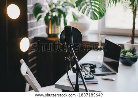 Items for recording podcast: professional microphone, earphones and laptop on white table in cozy home studio with black walls and lots of plants.