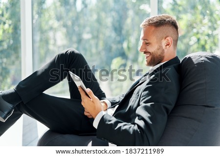 Smiling successful businessman in elegant suite using tablet sitting on bean bag chair with panoramic windows background in modern creative office