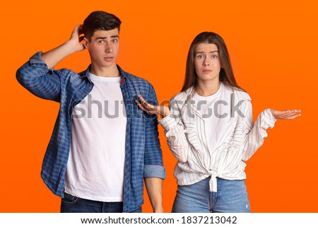 Puzzled young couple do not understand what happening or does something wrong. Surprised man scratches back of his head, sad woman spreads her arms to sides isolated on orange background, free space Royalty-Free Stock Photo #1837213042