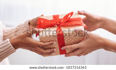 Holiday Celebration Concept. Closeup on female hands holding gift with red bow and giving present box to african american woman, greeting with Birthday, Christmas and New year at home