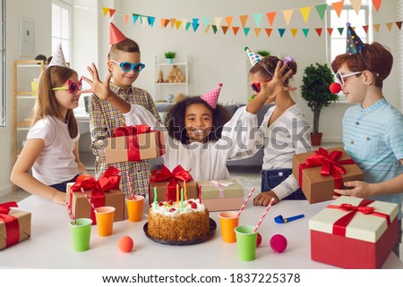 Happy African-American girl receiving birthday presents sitting at festive table with cake. Group of children in funny hats and sunglasses giving presents to their friend during fun party at home