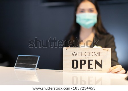 Business owner attractive young Asian woman in suitcase we're open sign on front counter welcoming clients to new hotel.Happy waiter with protective face mask holding open sign while stand at hotel .
