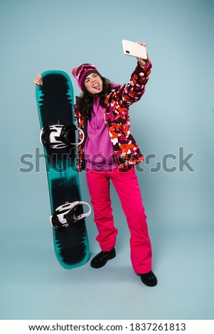 Joyful sportswoman showing her tongue while taking selfie with snowboard on cellphone isolated over blue wall