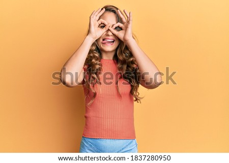 Young blonde girl wearing casual clothes doing ok gesture like binoculars sticking tongue out, eyes looking through fingers. crazy expression. 