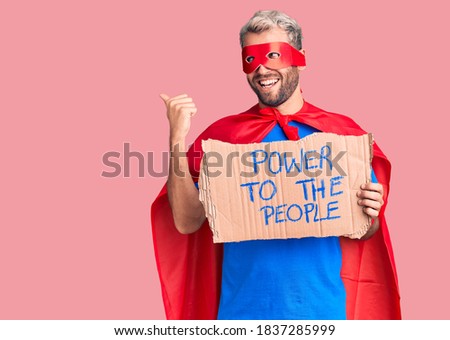 Young blond man wearing super hero custome holding power to the people cardboard banner pointing thumb up to the side smiling happy with open mouth 